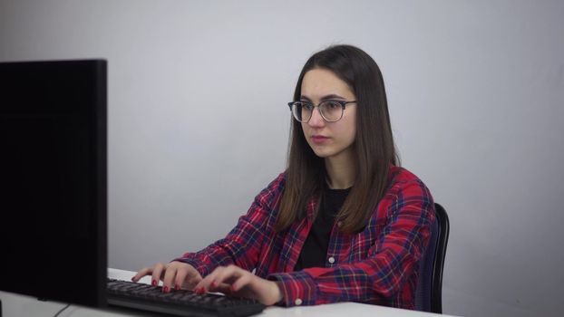 IT Specialist works for a PC. A young woman in glasses and a shirt is typing on a computer in the office.