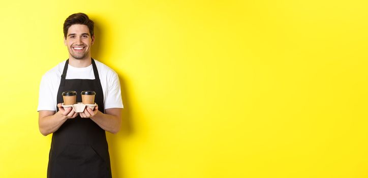 Handsome male barista serving takeaway coffee and smiling, bringing order, standing in black apron against yellow background