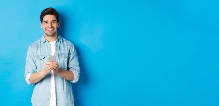 Handsome bearded man in casual outfit smiling at camera, checking smartphone, standing against blue background