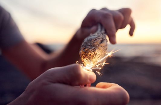 Hand, flame and sage burning at the beach for a spiritual ritual and meditation practice. Herb, finger and lighter with a plant being burned for zen meditative, mental health and chakra ceremony