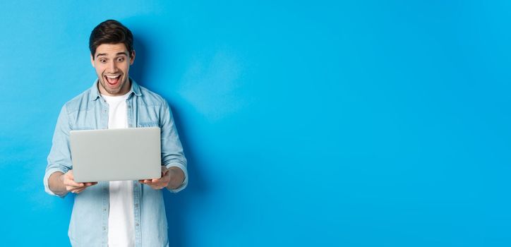 Image of amazed and happy man reacting to special offer in internet, looking at laptop excited, standing against blue background