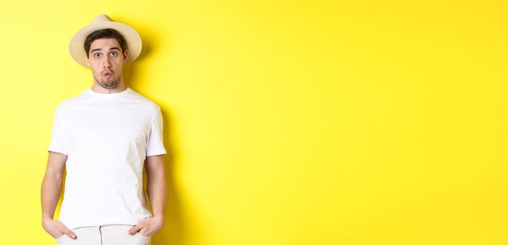 Confused guy traveller in straw hat looking puzzled, stare at camera, standing over yellow background