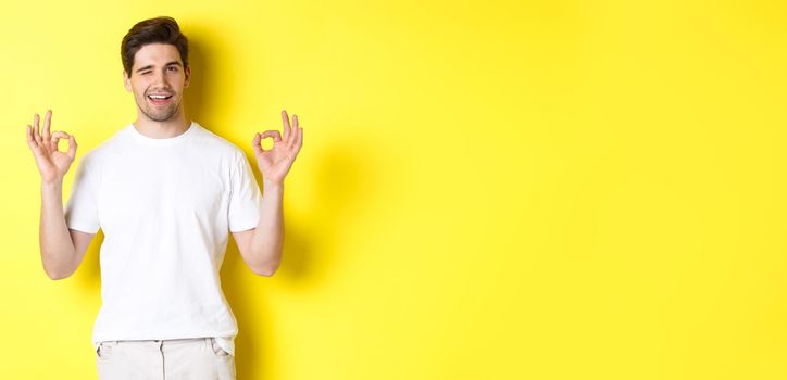 Confident handsome man winking, showing okay signs in approval, like something good, standing over yellow background