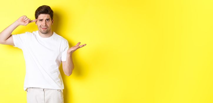 Confused and angry man pointing at head, scolding person for acting stupid, show crazy sign, standing over yellow background