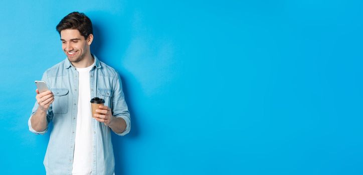 Handsome modern guy reading message on cell phone and drinking coffee, standing against blue background