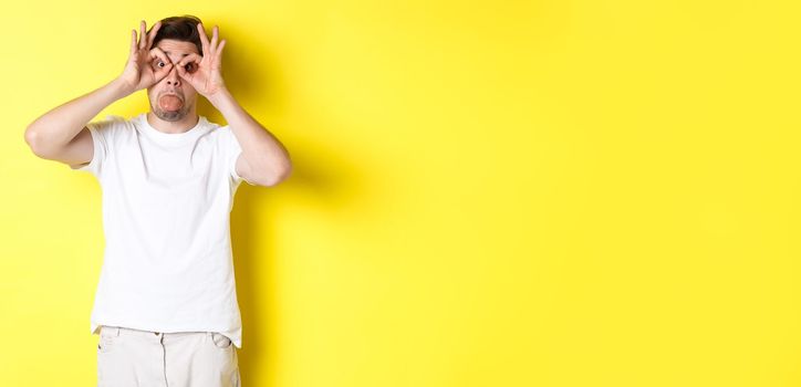 Young man making funny faces and showing tongue, fool around, standing in white t-shirt against yellow background