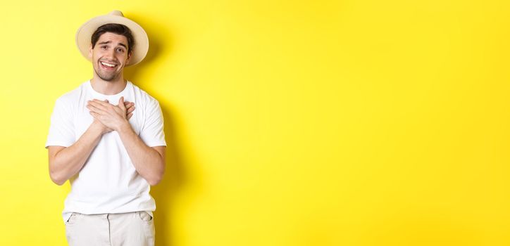 Concept of tourism and summer. Grateful guy in straw hat holding hands on heart, saying thank you and smiling with gratitude, standing against yellow background