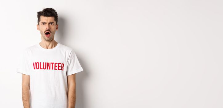 Shocked and confused man in volunteer t-shirt staring at camera and frowning displeased, standing against white background