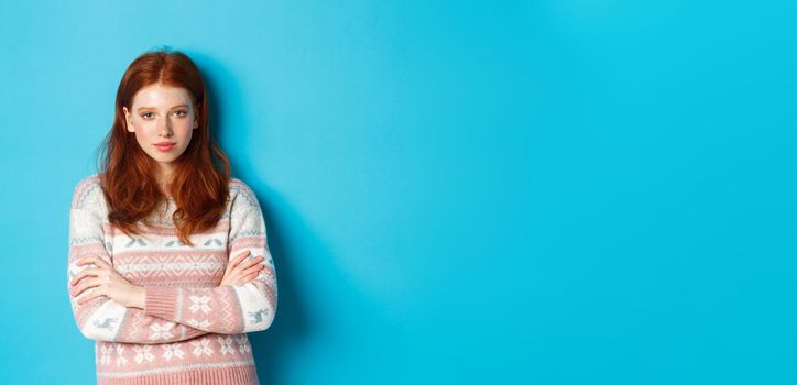 Image of confident teenage girl with red hair looking daring at camera, cross arms on chest and staring at camera, standing in winter sweater against blue background