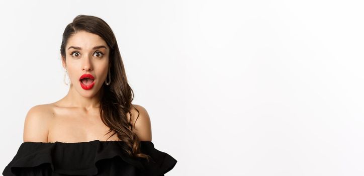 Fashion and beauty concept. Close-up of attractive brunette woman in black dress open mouth surprised, looking in awe at camera, white background
