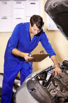 Step 1, check. A male mechanic holding a clipboard while working on the engine of a car.