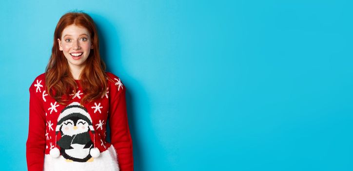 Winter holidays and Christmas Eve concept. Excited redhead girl in xmas sweater looking surprised at camera, standing against blue background