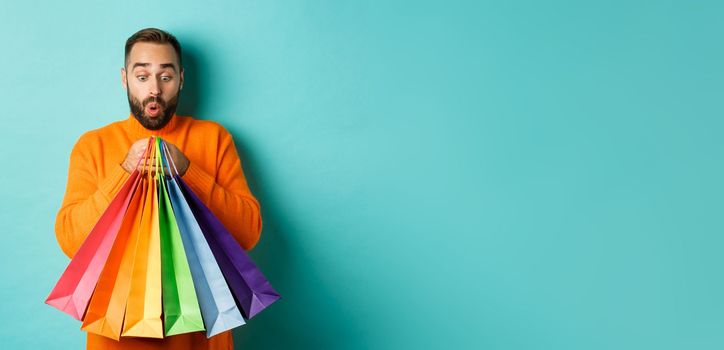 Excited man looking at shopping bags with purchases, standing amused against turquoise background