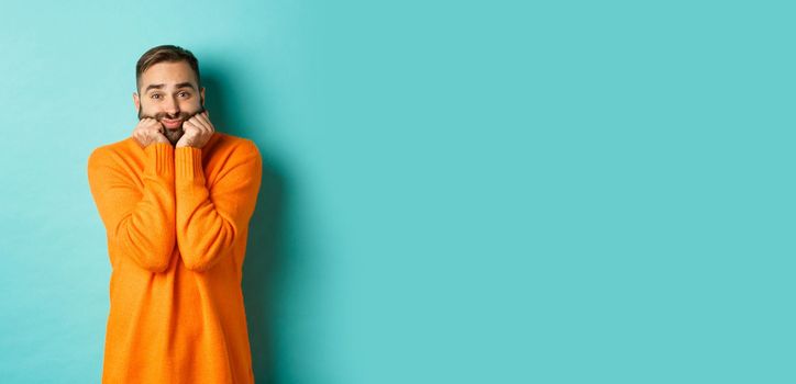 Sad man sighing and looking thoughtful, standing in orange sweater against turquoise background