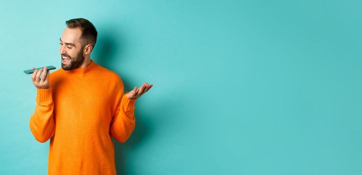 Happy man talking on speakerphone, gesturing and recording voice message on mobile phone, standing in orange sweater over light blue background