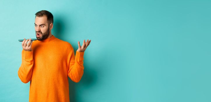 Angry man arguing on speakerphone, record voice message with mad face, standing over light blue background in orange sweater