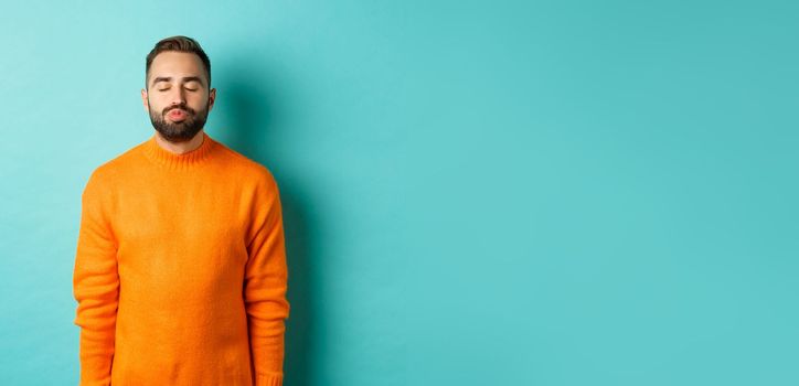 Waist up shot of young man pucker lips and close eyes, waiting for kiss, standing in orange sweater against light blue background