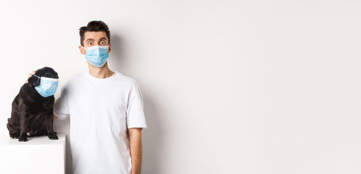 Covid-19, animals and quarantine concept. Image of funny young man and pug in medical masks, staring at camera, standing over white background