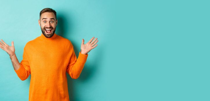 Excited man react to promo offer, spread hands sideways and rejoicing, standing against turquoise background in orange sweater