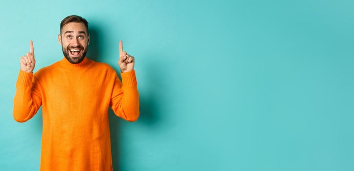 Happy caucasian man showing winter holidays promo offer, pointing fingers up, demonstrate advertisement, standing in sweater against turquoise background