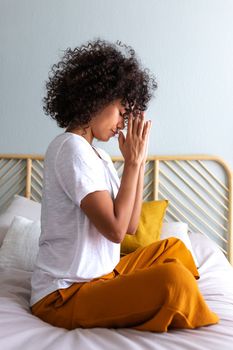 Side view of spiritual young multiracial woman meditating with hands in prayer at home sitting on bed. Vertical