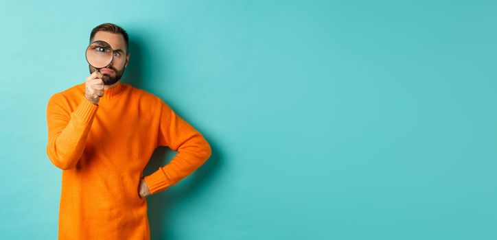 Funny man looking serious through magnifying glass, inspecting something, standing in orange sweater against turquoise background