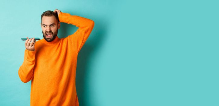 Confused man scratching head while talking on speakerphone, record voice message with indecisive face, standing in orange sweater over light blue background