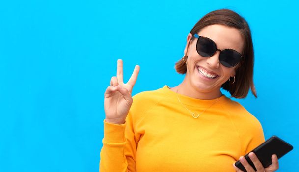 Im all about peace and optimism. Cropped portrait of a happy young woman showing the peace sign against a blue background.