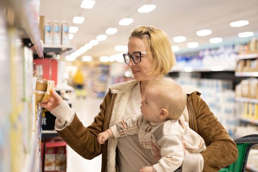 Caucasian mother shopping with her infant baby boy child choosing products in department of supermarket grocery store.