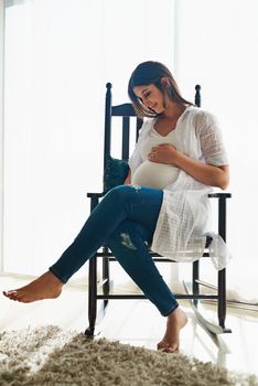 I cant wait to meet you. a happy pregnant woman cradling her belly while relaxing in a chair at home.