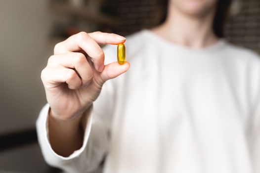 woman's hand holds an Omega-3 capsule against the background of herself in a white sweater, the background is in blur. Useful fish oil capsule tablets