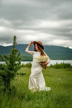 A middle-aged woman in a white dress and brown hat holds a lA middle-aged woman in a white dress and brown hat holds a basket in her hands with a large bouquet of daisies.arge bouquet of daisies in her hands. Wildflowers for congratulations