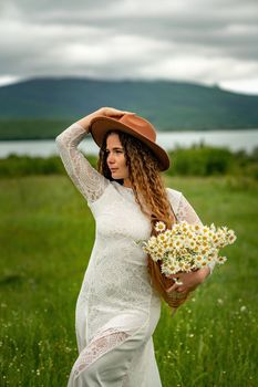 A middle-aged woman in a white dress and brown hat holds a lA middle-aged woman in a white dress and brown hat holds a basket in her hands with a large bouquet of daisies.arge bouquet of daisies in her hands. Wildflowers for congratulations