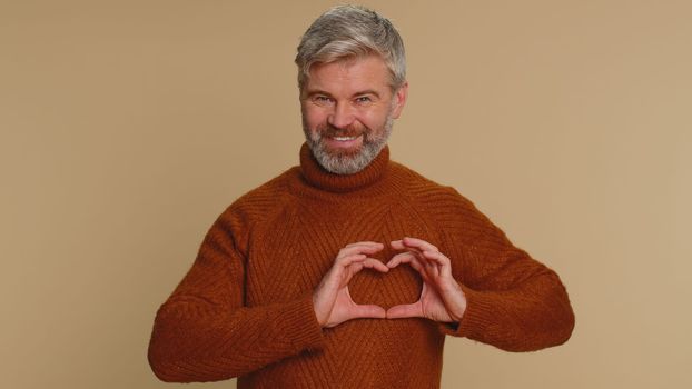 Smiling mature man makes heart gesture demonstrates love sign expresses good feelings and sympathy