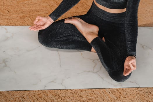 Close-up of the legs and arms of a woman in a black tracksuit sitting on a yoga mat in a lotus position in the gym