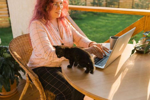 Woman working with laptop outdoor in summer terrace with black kitten. Girl and cat watching video on computer in the evening. Working from home, business, pet and lifestyle concept.