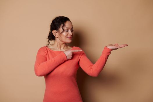 Gorgeous Hispanic woman points at copy advertising space on her hand palm up, isolated on color beige background. Mockup
