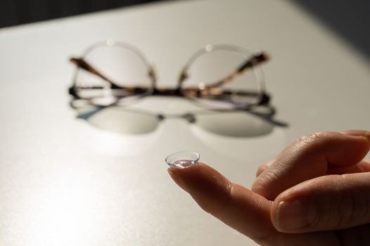 Close-up of a contact lens on a female index finger against the background of glasses on a white table.