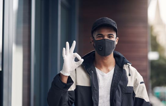 Precautionary measures and professionalism go hand in hand. a masked and gloved young man showing an okay sign while making a home delivery.