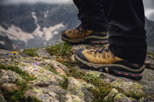close-up, legs in trekking boots stand on a stone near a cliff against the backdrop of snow-capped mountains. hike
