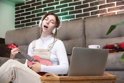 young woman student learning vocals and playing the ukulele at home sitting on the floor in the living room in front of a laptop