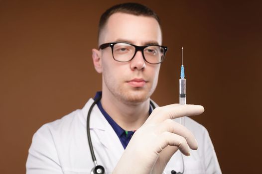Caucasian male doctor holding a syringe in a bathrobe with a stethoscope and gloves on a plain background, pensive look, studio shot