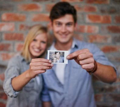 Creating memories. A young couple standing in front of a brick wall holding a photo to the camera.