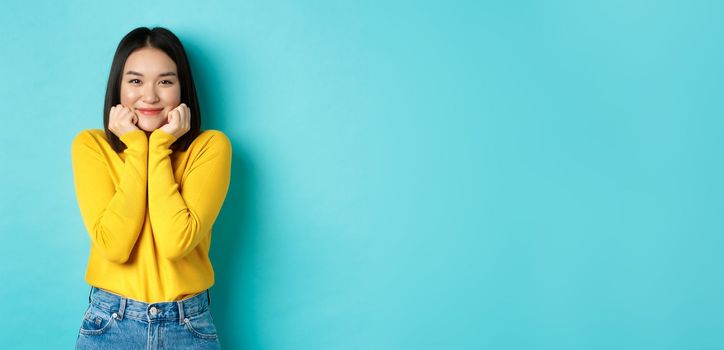 Beauty and fashion concept. Beautiful asian woman blushing and smiling, looking at something cute and silly, lean face on palms, standing over blue background