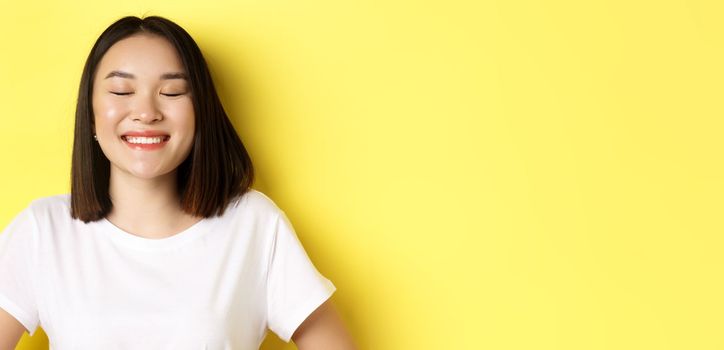 Close up of happy, romantic asian girl dreaming of something, close eyes and smiling delighted, standing over yellow background