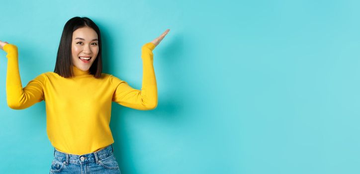 Beauty and fashion concept. Stylish beautiful asian girl demonstrate promotion, raising hands up and smiling, showing something on blue background.