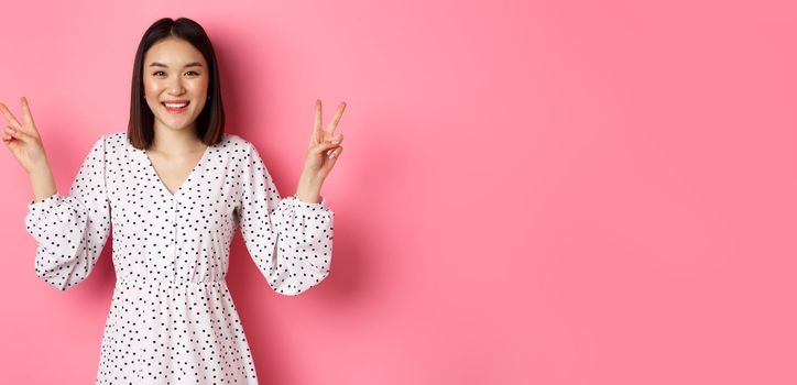 Cute asian brunette girl in dress smiling, showing kawaii peace signs and looking happy, standing over pink background
