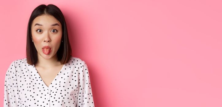 Beauty and lifestyle concept. Close-up of funny and cute asian woman showing tongue, staring at camera silly, standing over pink background