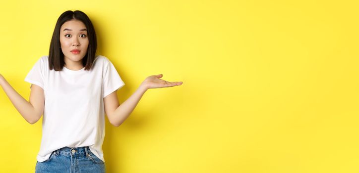 Clueless asian girl shrugging shoulders, spread out hands and looking confused, know nothing, standing over yellow background