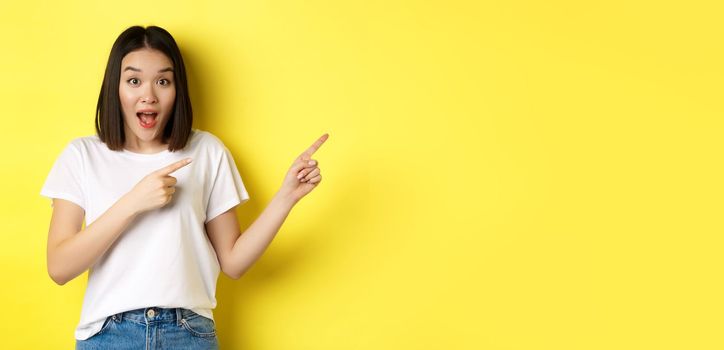 Beauty and fashion concept. Beautiful asian woman in white t-shirt pointing fingers right, demonstrate logo standing over yellow background